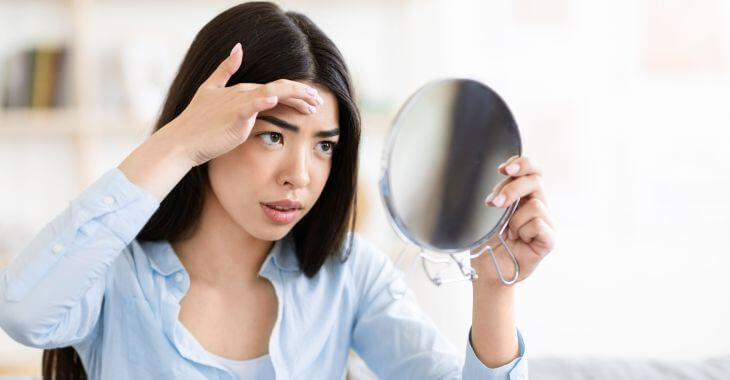 Concerned young woman looking at a growth on her forehead in the mirror.