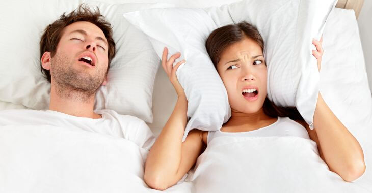 An annoyed woman covering her ears with a pillow due to her partner's snoring caused by sleep apnea.