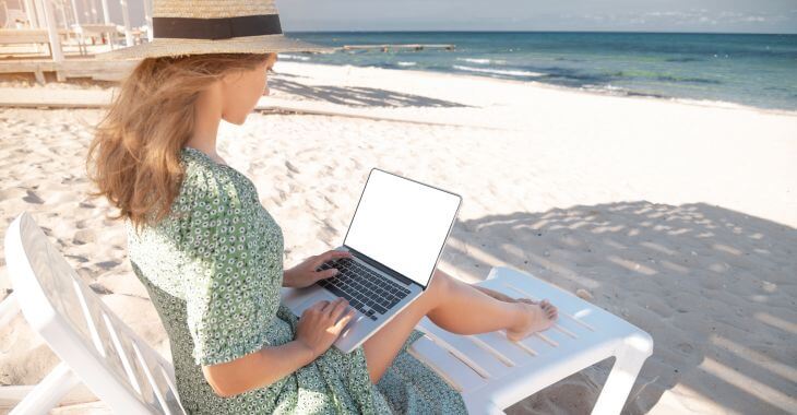 A woman working on a laptop on a beach hiding in the shade of the beach umbrella.