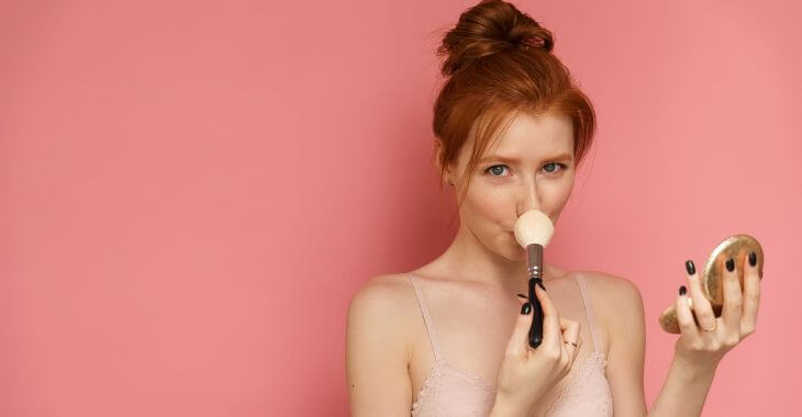 Red-haired slim woman covering her nose with a make-up brush.