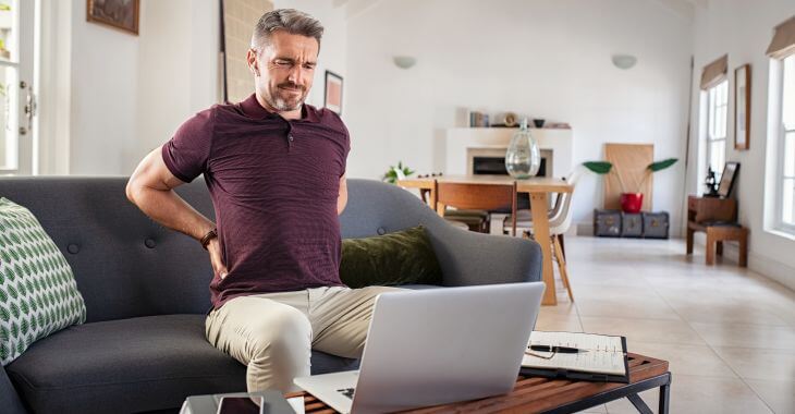A mature man with lumbar pain sitting on a couch in front of a laptop. 