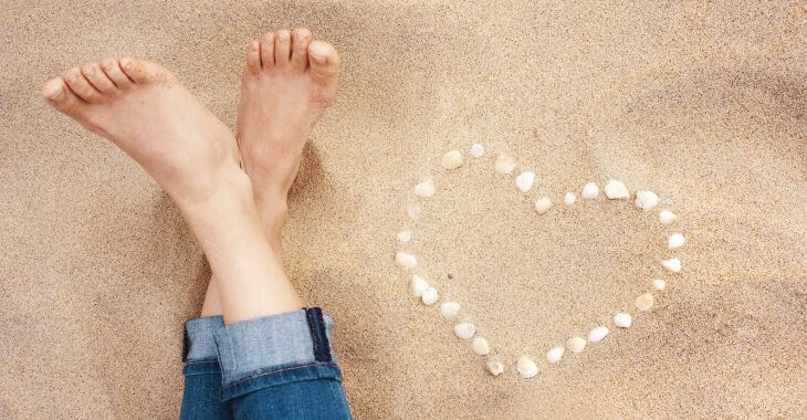 Woman's feet next to a heart made of a seashells on the beach.