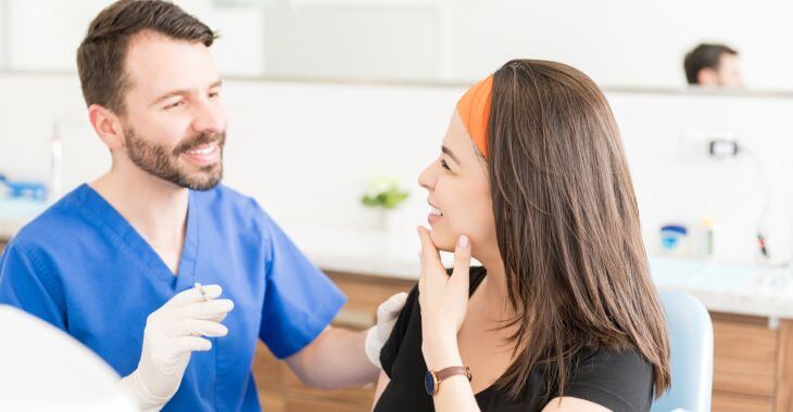 A cosmetic surgeon holding a syringe with filler talking to a happy woman touching her chin.