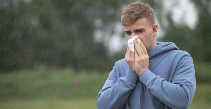 A teenage boy blowing his nose while walking through the park in the rain. 