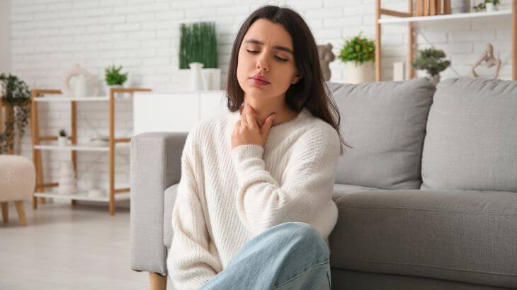 An upset young woman suffering from voice and throat problems siting on a floor near the couch and rubbing her neck 