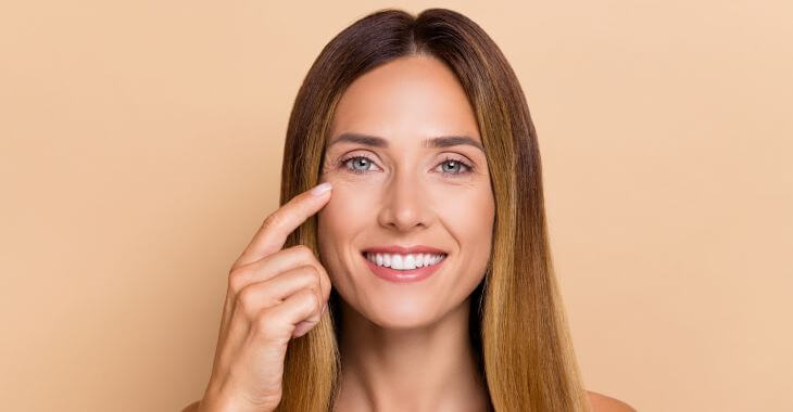 Youngish looking middle-aged woman after undereye rejuvenation pointing to her skin under eyes