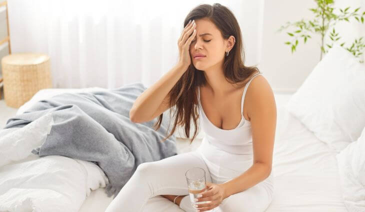 A woman with headache sitting on the bed after waking up in the morning and holding a glass of water