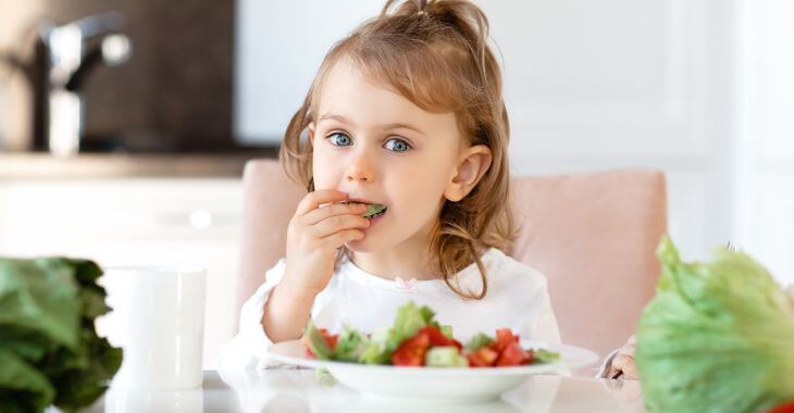 A little girl sitting at a table over a plate with a salad and eating vegies 