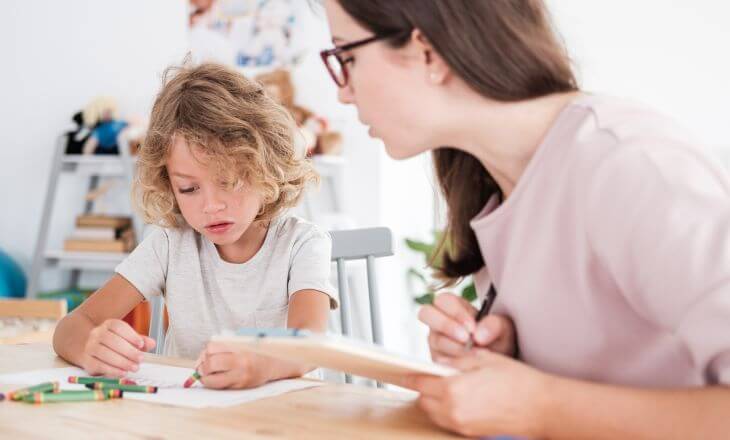 A pediatric speech theraphist sitting next to a kid who is drawing a picture 