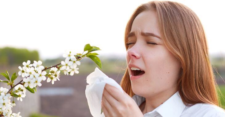 Sneezing young woman suffering from allergy standing near blooming tree