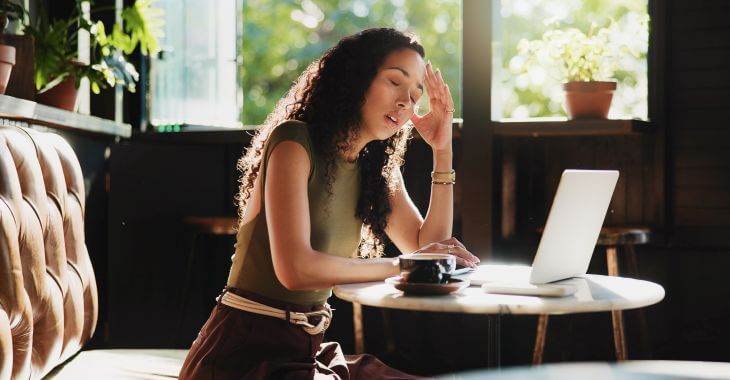 A tirede sleepy woman sitting at the cafe table with an open laptop and a cup of coffee